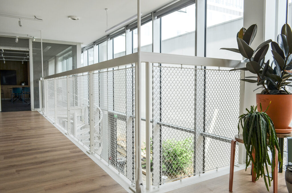 Una oficina moderna con una pared de vidrio de dos pisos en la entrada, que permite que la luz natural inunde el espacio. Una barandilla de metal blanco está elegantemente instalada a lo largo del pasillo del nivel superior, lo que brinda seguridad e iluminación adicional. Las barandillas de malla de acero inoxidable Gaudi F de Codina mejoran el diseño interior de la oficina al mismo tiempo que promueven la seguridad y la estética.